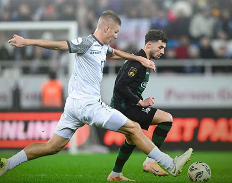 Два футболиста борются за мяч на поле во время матча. Игрок в белой форме вытягивает ногу для отбора, противостоя игроку в черной форме. На заднем плане видны трибуны с болельщиками